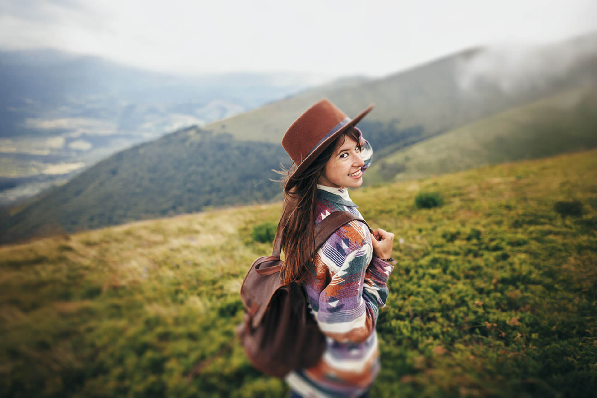 Junge Person mit Hut und Rucksack blickt lächelnd beim Wandern über die grünen Hügel des Allgäus
