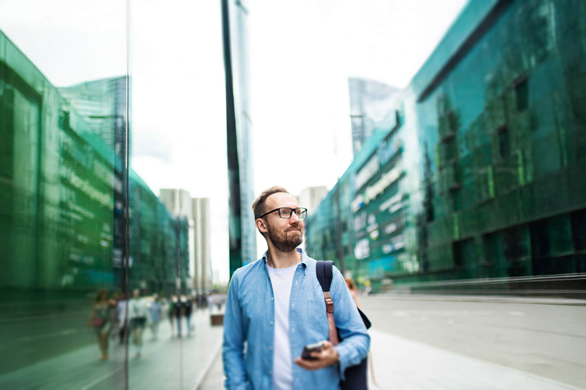 Junger Mann mit Brille und Smartphone geht durch eine moderne Glasfassaden-Straße in Newcastle auf dem Weg zur Sprachschule.