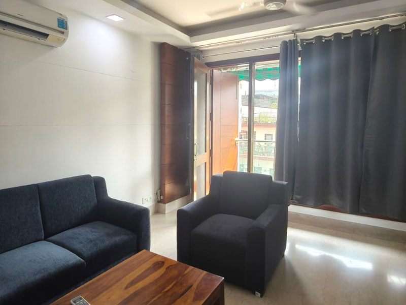 Helles Wohnzimmer im Apartment in Neu-Delhi mit Sofa, Sessel und Zugang zum Balkon.