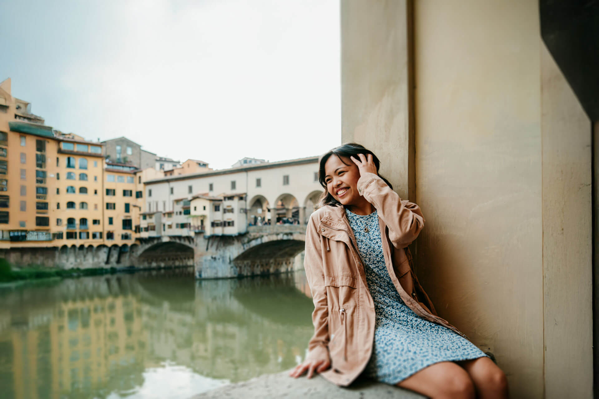 Lächelnde junge Frau sitzt am Fluss Arno mit Blick auf die Ponte Vecchio in Florenz