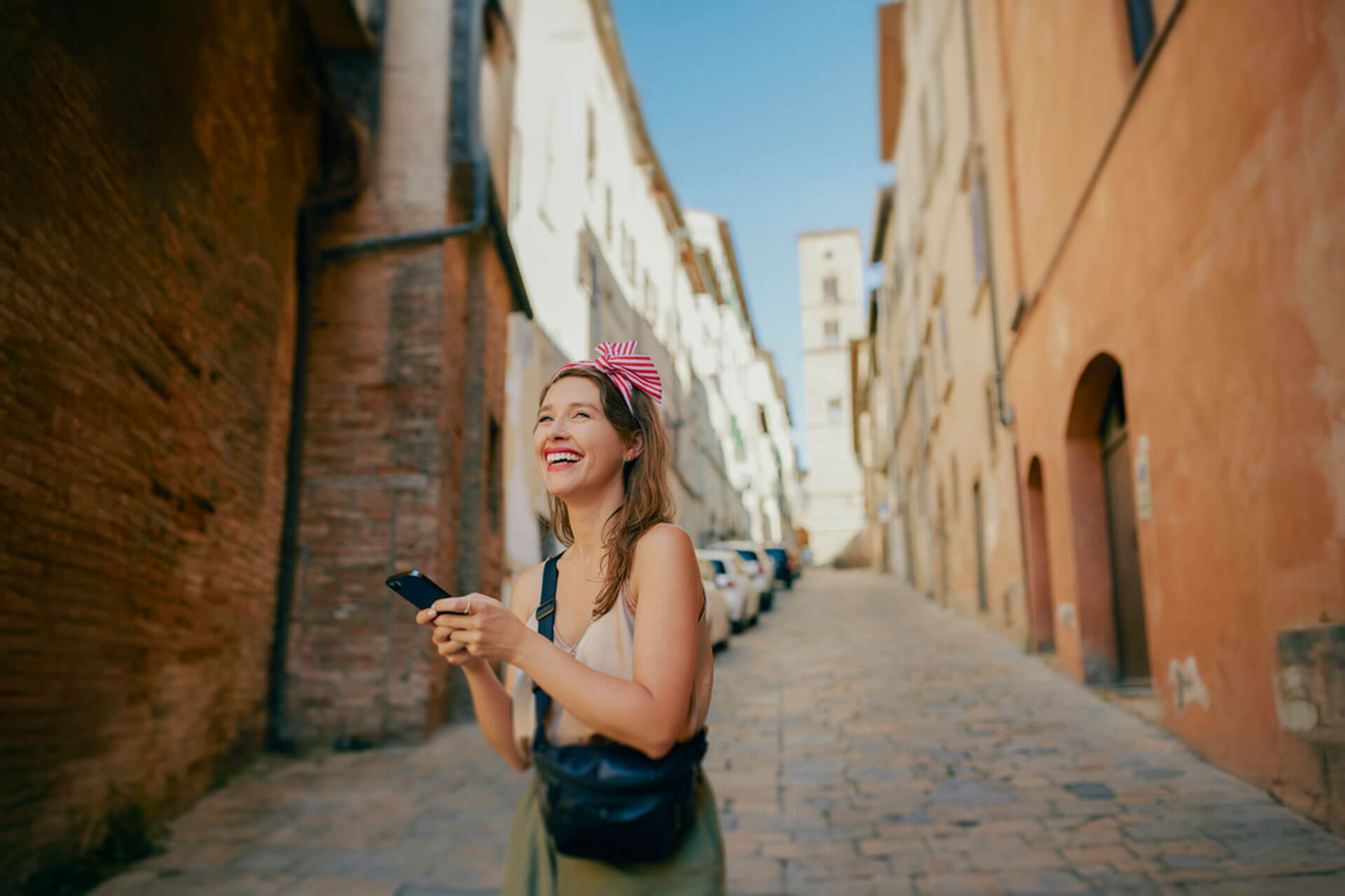 Junge Frau lacht fröhlich und hält ihr Smartphone in einer sonnigen Gasse in Siena