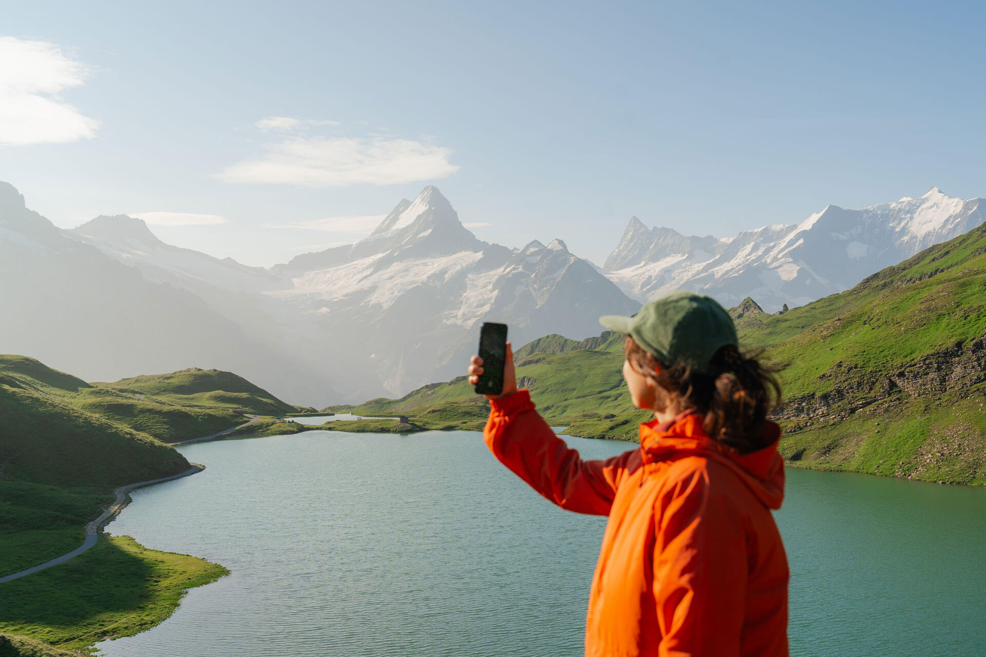 Person in oranger Jacke fotografiert die Berglandschaft mit See und schneebedeckten Gipfeln in der Schweiz