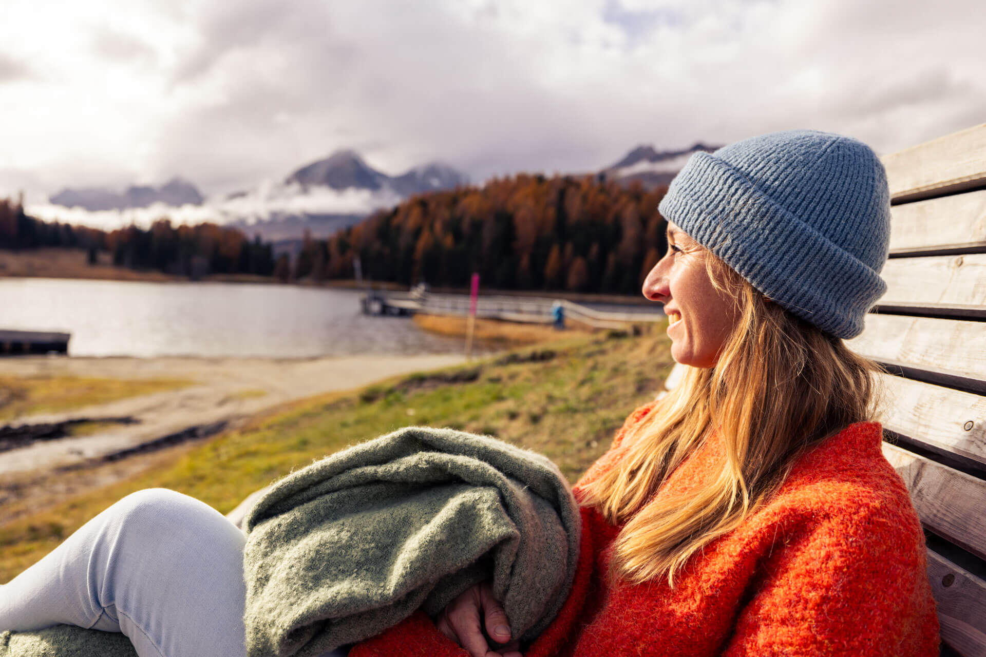 Person in warmer Kleidung entspannt auf einer Bank mit Blick auf See und Alpenlandschaft