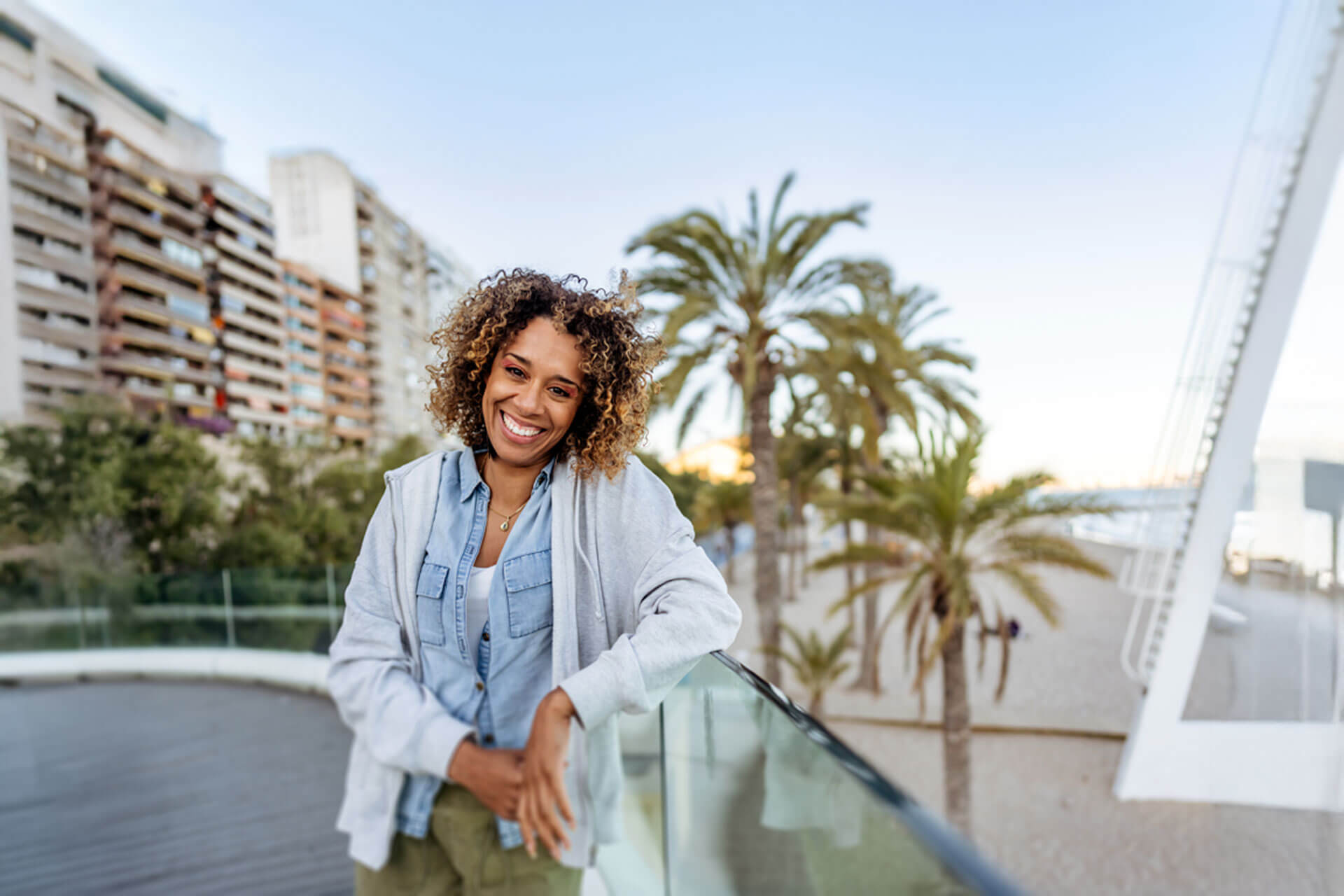 Frau lehnt sich lächelnd an eine Glasbrüstung mit Palmen und Skyline im Hintergrund von Miami