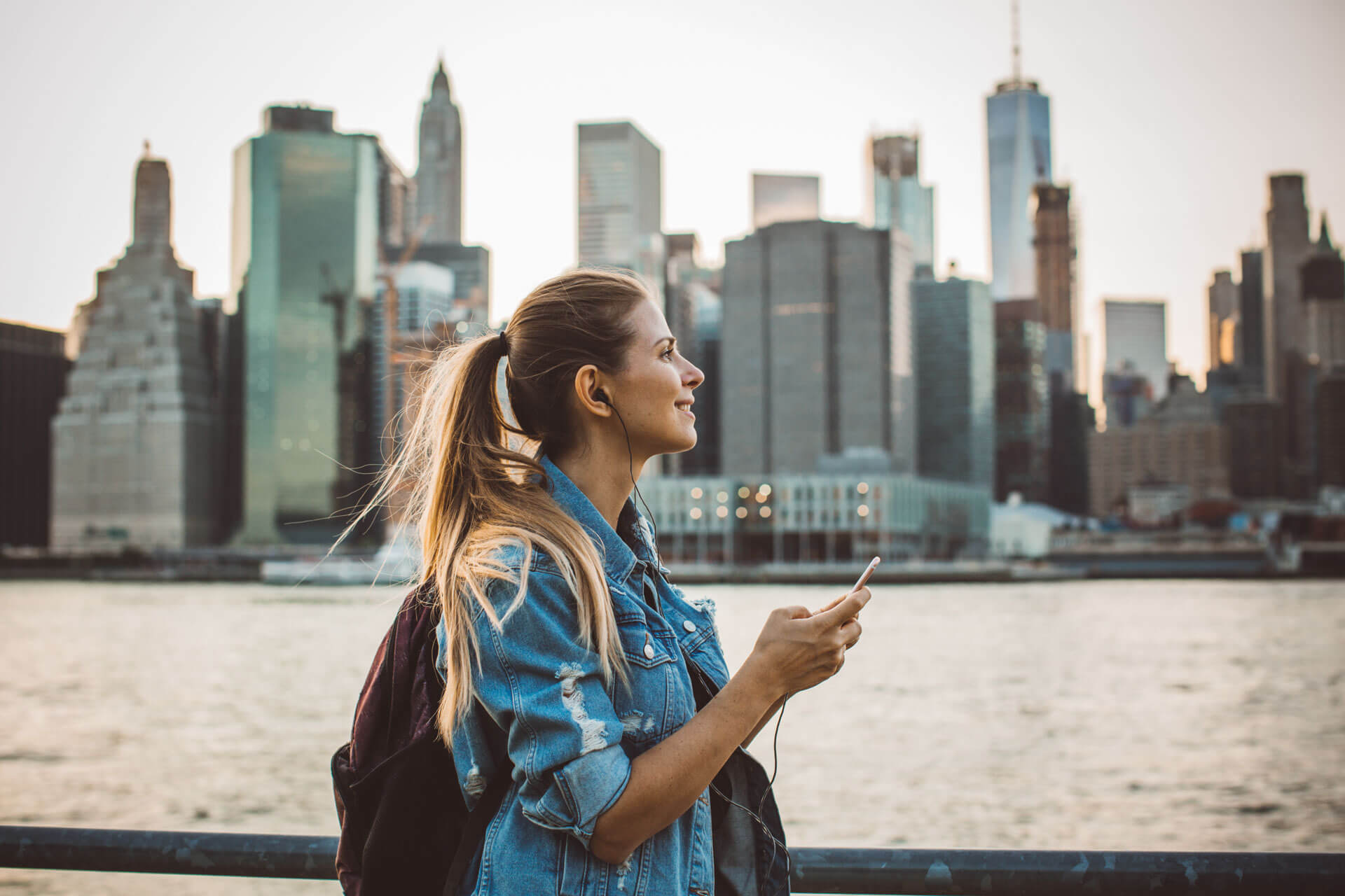 Frau mit Kopfhörern blickt lächelnd auf die Skyline von Manhattan in New York
