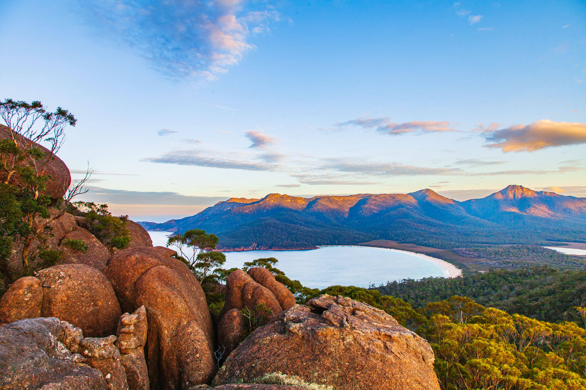 Aussicht auf die Wineglass Bay in Tasmanien mit roten Felsen im Vordergrund und Bergen im Hintergrund