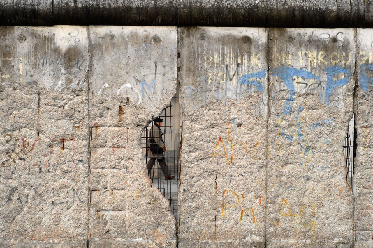 Erhaltener Abschnitt der Berliner Mauer mit Ausbruchstelle; Person läuft dahinter vorbei.