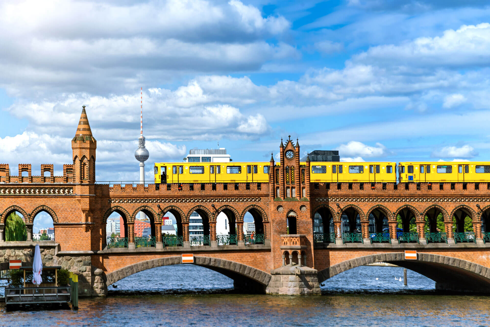 Gelbe U-Bahn über die Oberbaumbrücke, Spree darunter, Fernsehturm im Hintergrund bei blauem Himmel.