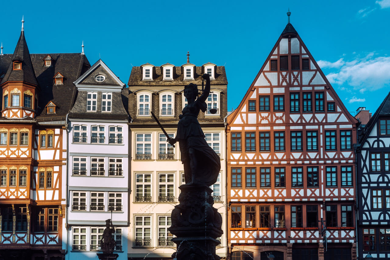 Fachwerkhäuser am Römerberg mit Justitia-Statue im Vordergrund vor blauem Himmel.