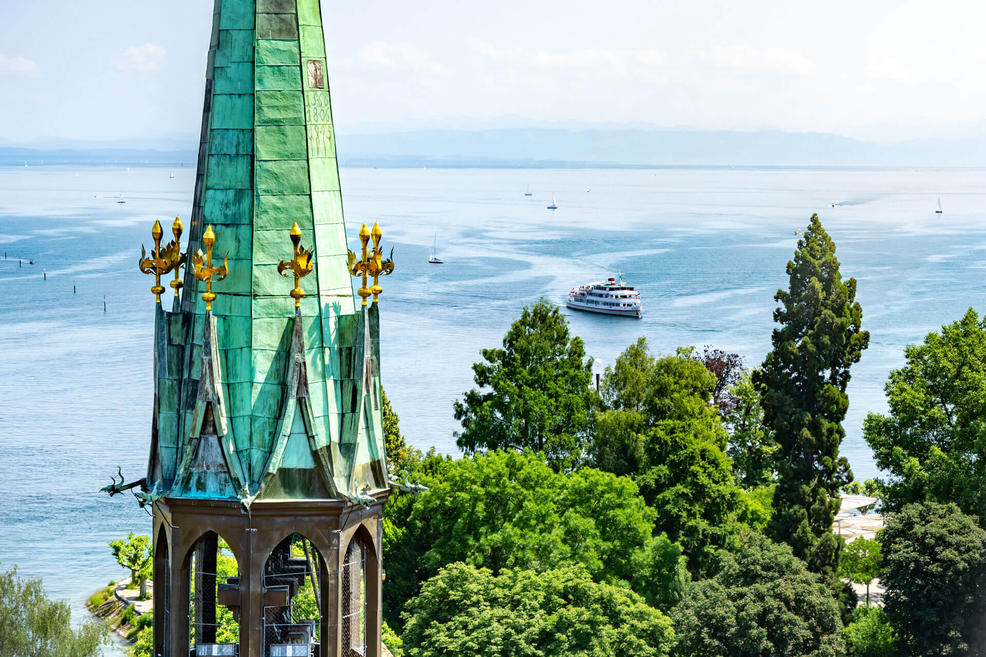 Historischer Turm mit grünem Kupferdach am Bodensee, Ausflugsschiff und Alpenpanorama im Hintergrund