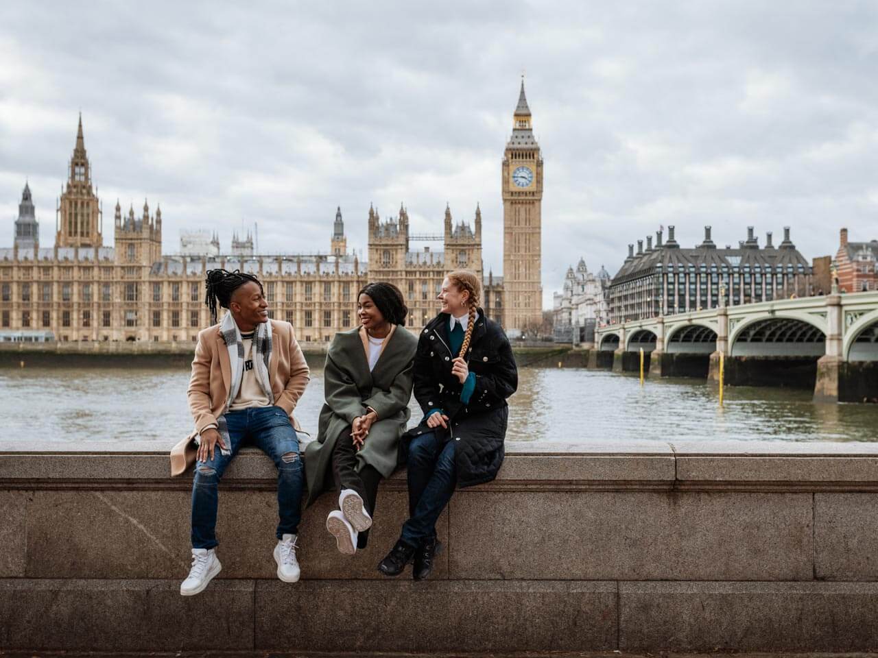 Drei junge Menschen sitzen an der Themse vor Big Ben und Westminster und plaudern Small Talk und Redewendungen in authentischen Gesprächen trainieren