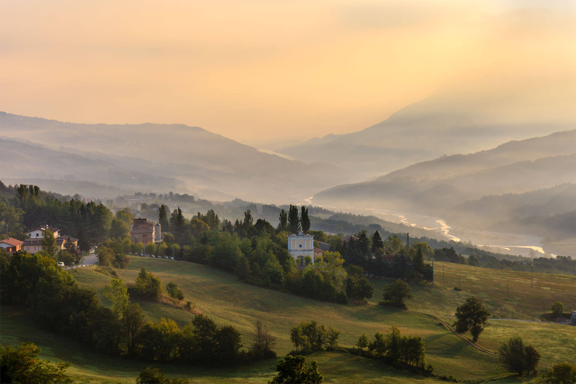 Sonnenaufgang über den nebligen Hügeln bei Bagno di Romagna, mit grünen Wiesen und verstreuten Häusern im Vordergrund