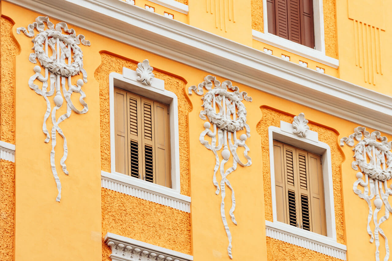 Detail einer verzierten Hausfassade in Triest mit leuchtend gelber Farbe und weißen Stuckornamenten, Sprachkurs in Italien mit Kulturbezug.