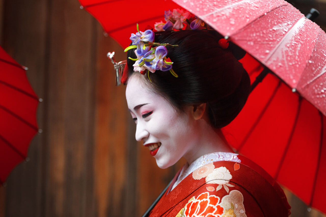 Maiko im roten Kimono unter rotem Schirm lächelt im Regen in Kyoto