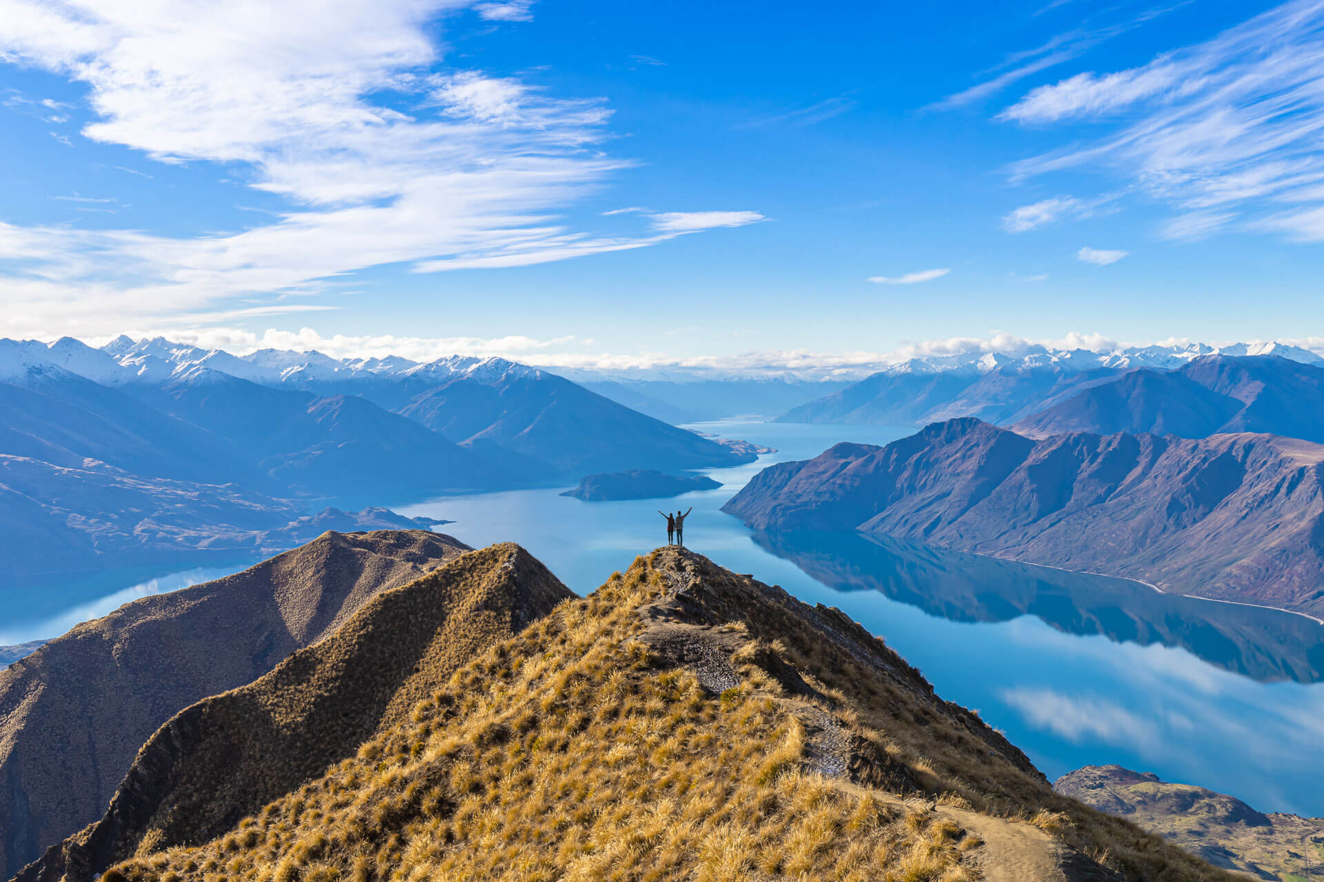 Zwei Wanderer stehen auf dem Roys Peak mit Blick auf den Lake Wanaka und die Berge Neuseelands.