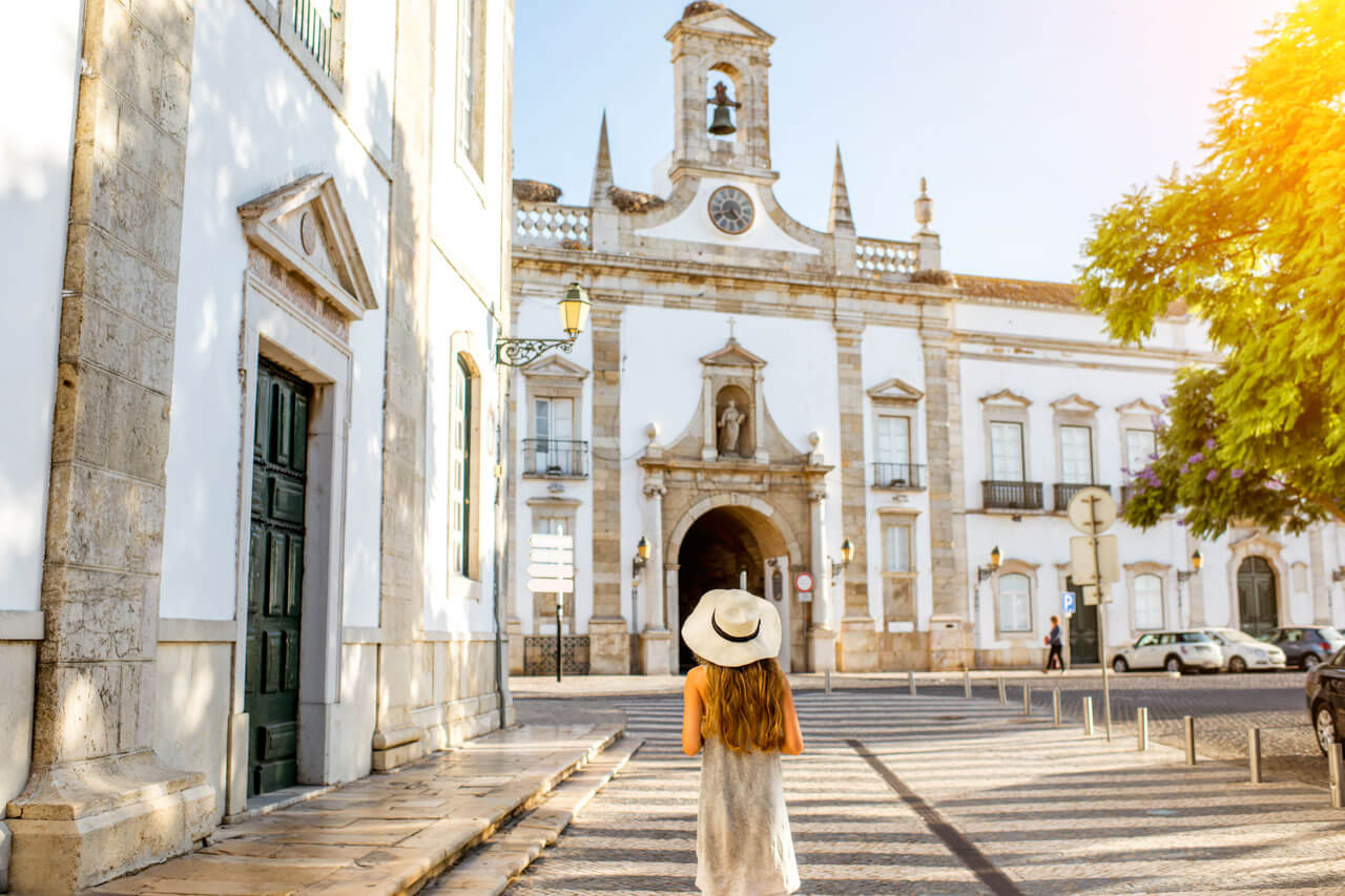 Frau mit Sommerhut spaziert durch die Altstadt von Faro und blickt auf das Arco da Vila – Sprachkurs in Portugal.