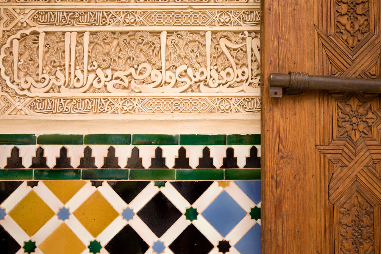 Alhambra-Detail: arabische Stuckinschrift über farbigen Kacheln neben geschnitzter Holztür.