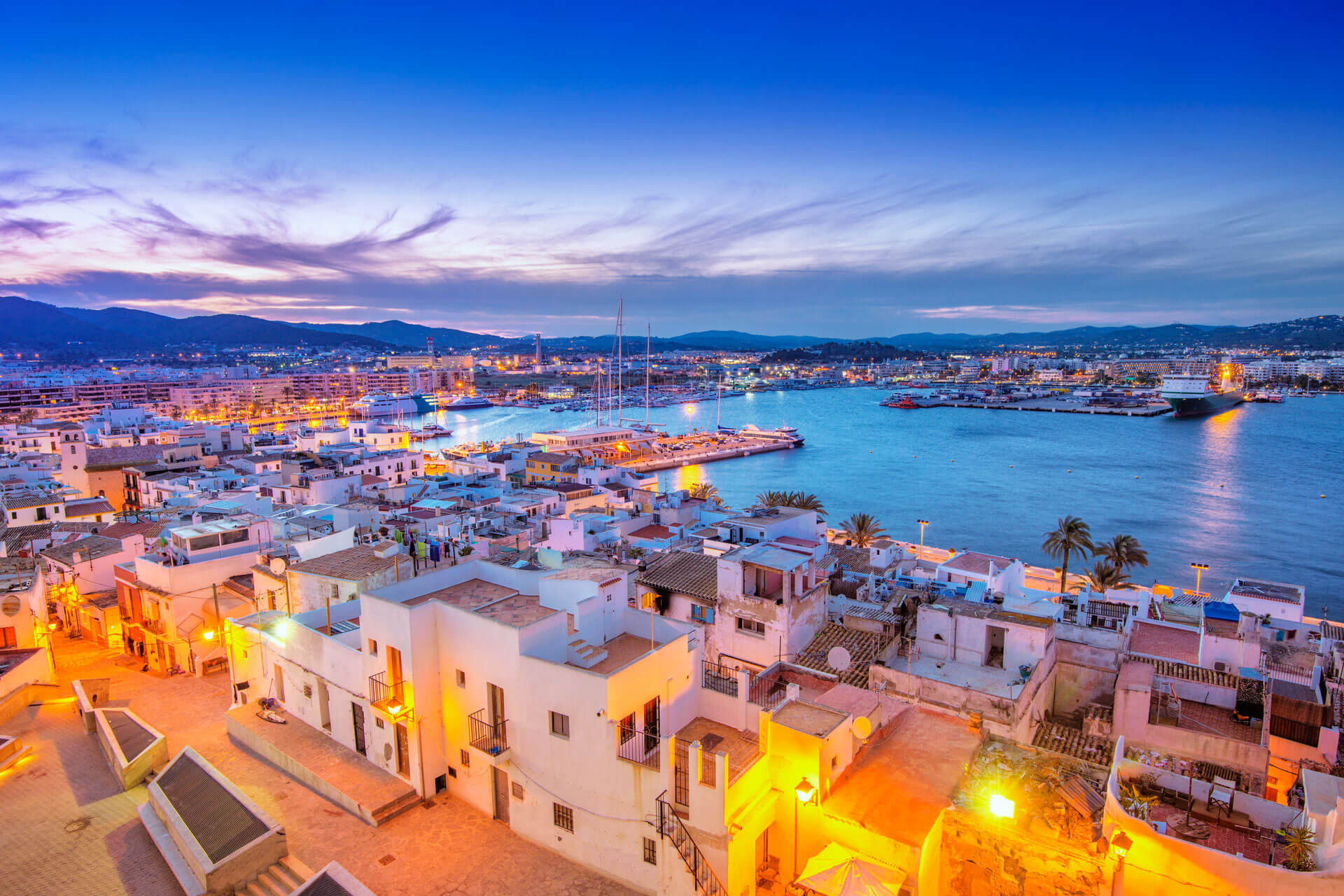 Abendblick über Ibiza-Stadt (Eivissa) und den Yachthafen in der blauen Stunde – warm beleuchtete weiße Häuser am Meer.