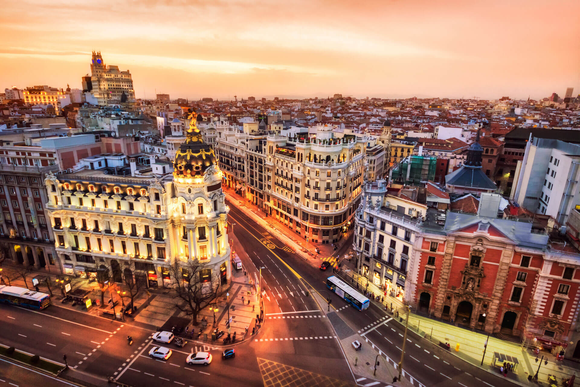 Luftaufnahme der Gran Vía in Madrid bei Sonnenuntergang mit dem beleuchteten Metropolis-Gebäude und Lichtspuren des Verkehrs.