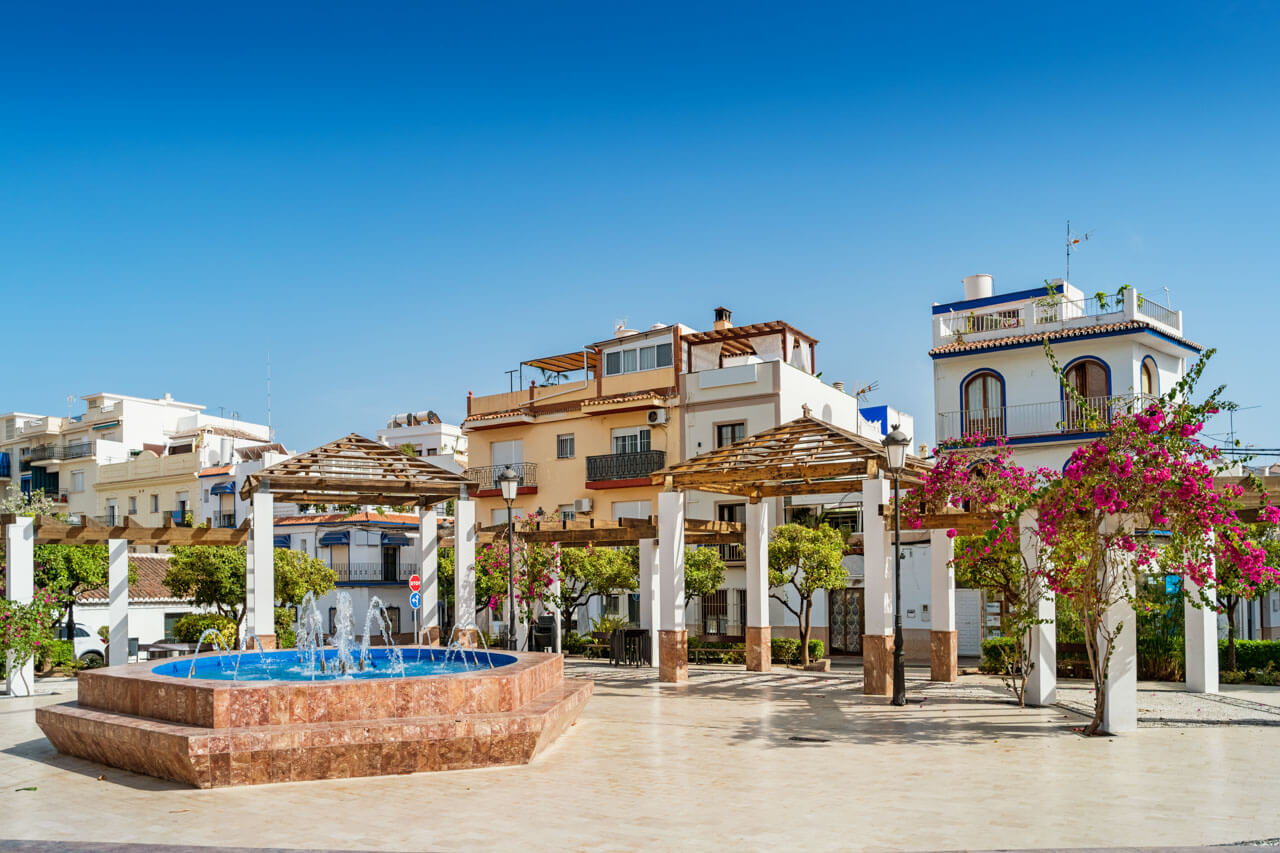 Sonniger Platz in Nerja mit Springbrunnen, Pergolen und weiß-blauen Häusern