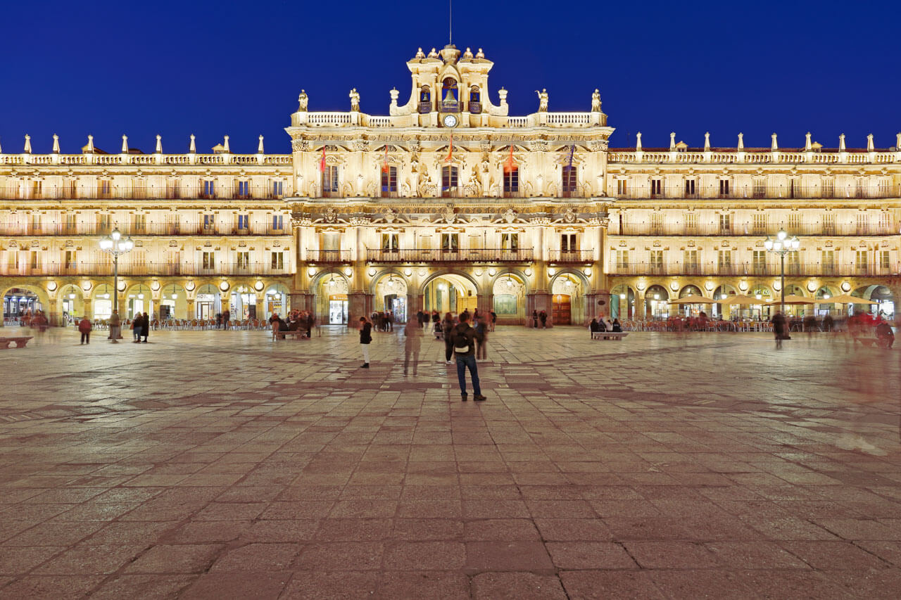 Beleuchtete Plaza Mayor in Salamanca bei Nacht mit barocken Arkaden und Menschen auf dem weiten Platz.