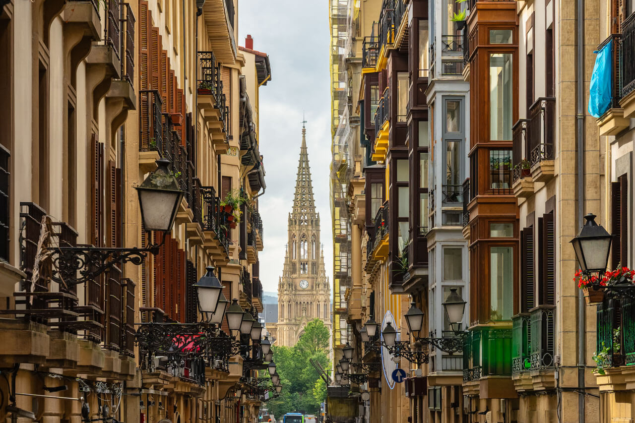 Enge Altstadtgasse mit schmiedeeisernen Balkonen; am Ende ragt die Turmspitze der Kathedrale Buen Pastor.