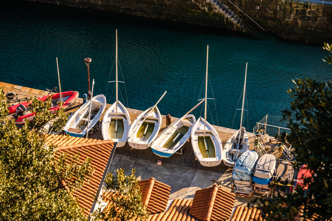 Kleine Segeljollen am Kai des Hafens von San Sebastián neben türkisblauem Wasser und Ziegeldächern.