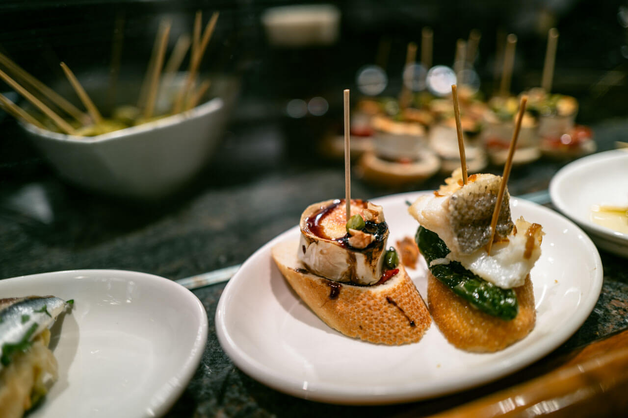 Pintxos auf Baguettescheiben – Ziegenkäse mit Balsamico sowie Kabeljau mit Padrón-Paprika auf einem Teller.
