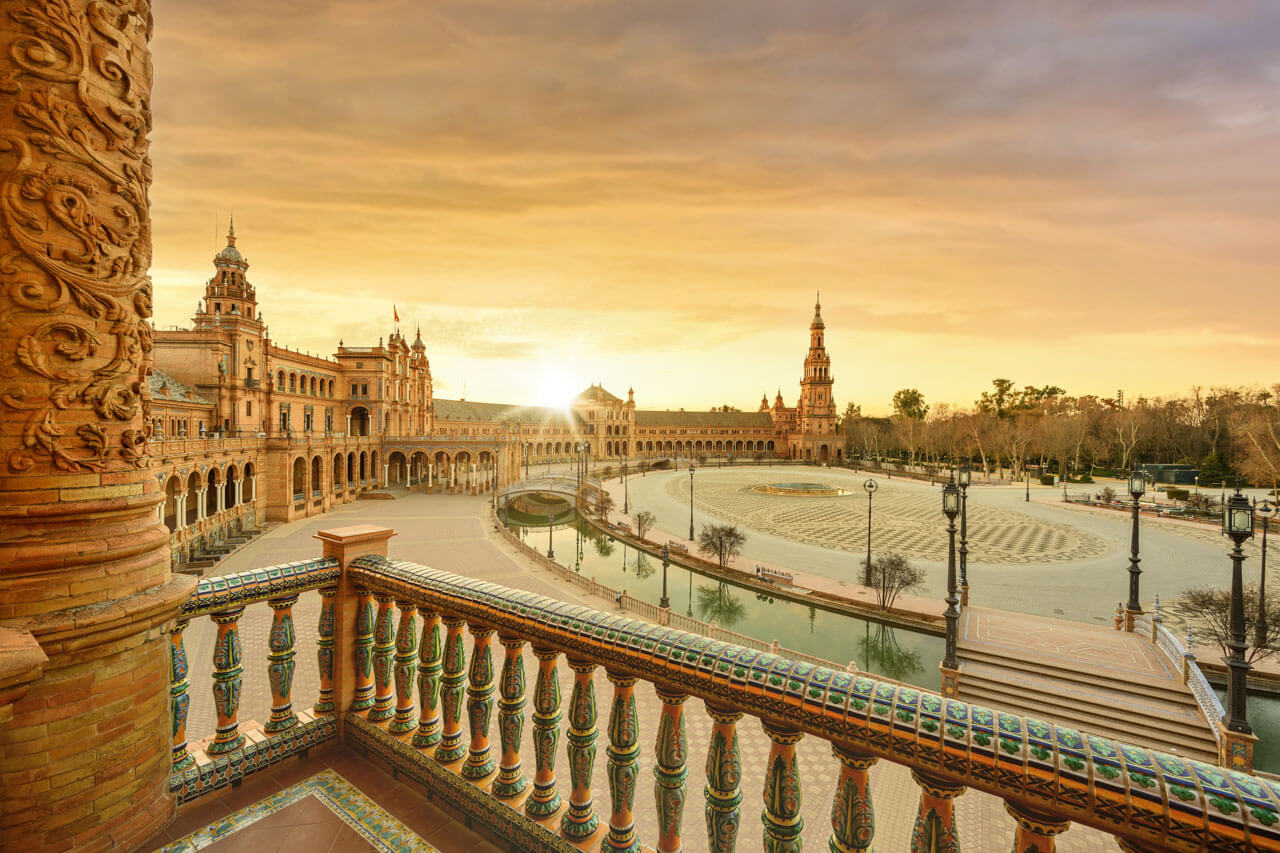 Panoramablick bei Sonnenaufgang über die Plaza de España in Sevilla – vom bunt gekachelten Balkon auf Bögen, Türme, Kanal und Springbrunnen.