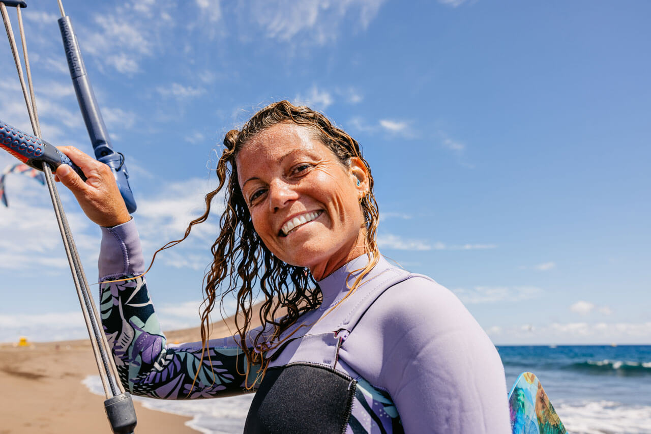 Lachende Kitesurferin im Neoprenanzug am Strand, nasses Haar, Kite-Bar in der Hand, Meer und blauer Himmel.