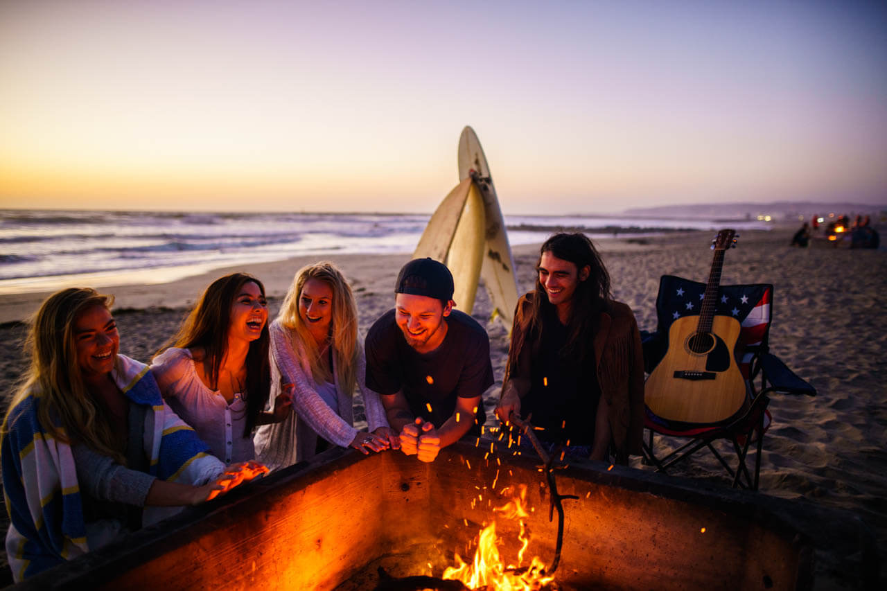 Freunde am Strandfeuer bei Sonnenuntergang, Gitarre und Surfbrett. Konversation üben im Abendlicht.