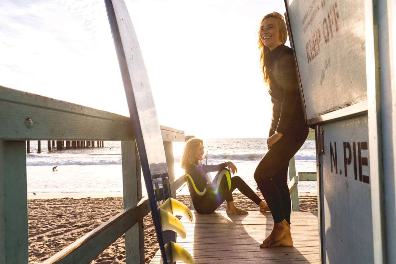 Zwei Surferinnen in Neoprenanzügen am Rettungsturm; Boards im Sand; Sonnenuntergang am Strand.