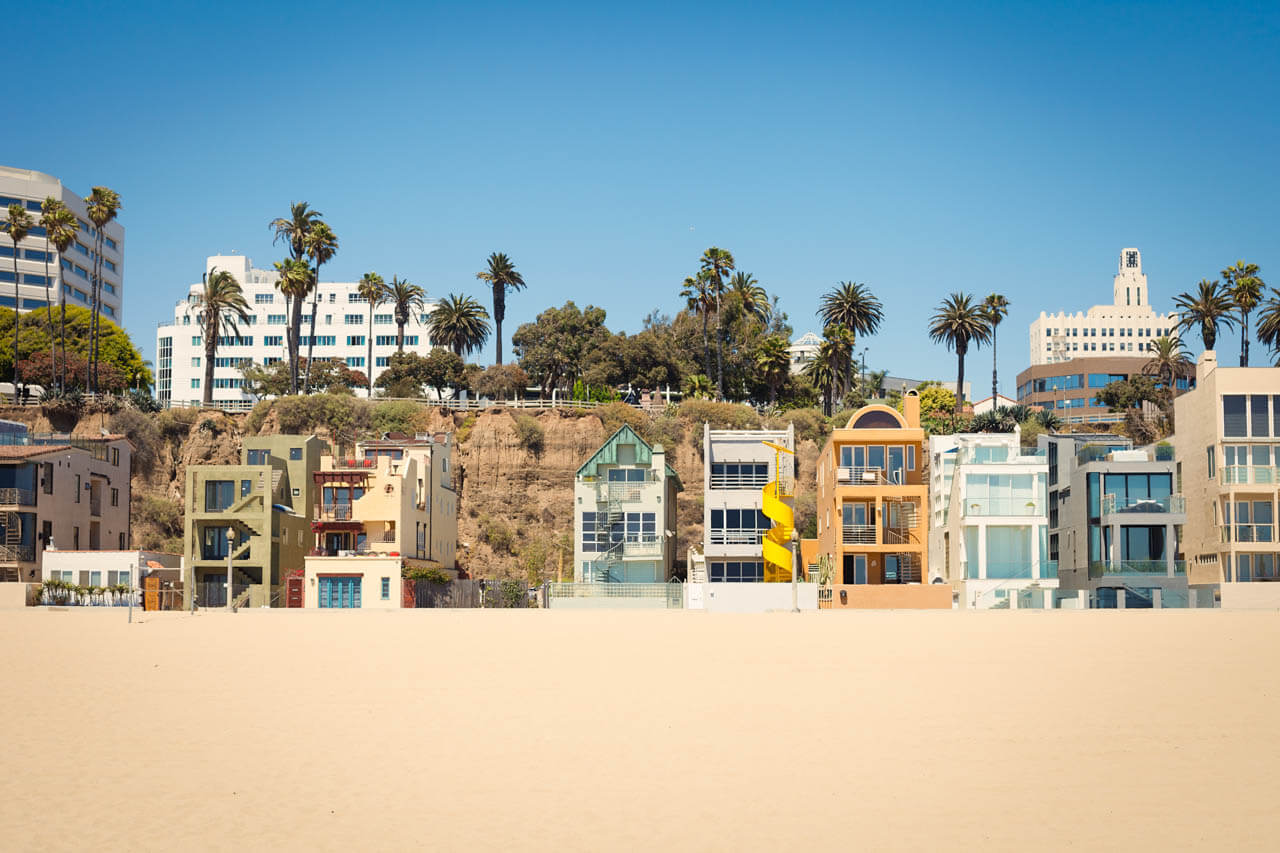 Bunte Strandhäuser und Palmen vor der Steilküste; klarer Himmel über dem weiten Strand von Santa Monica.